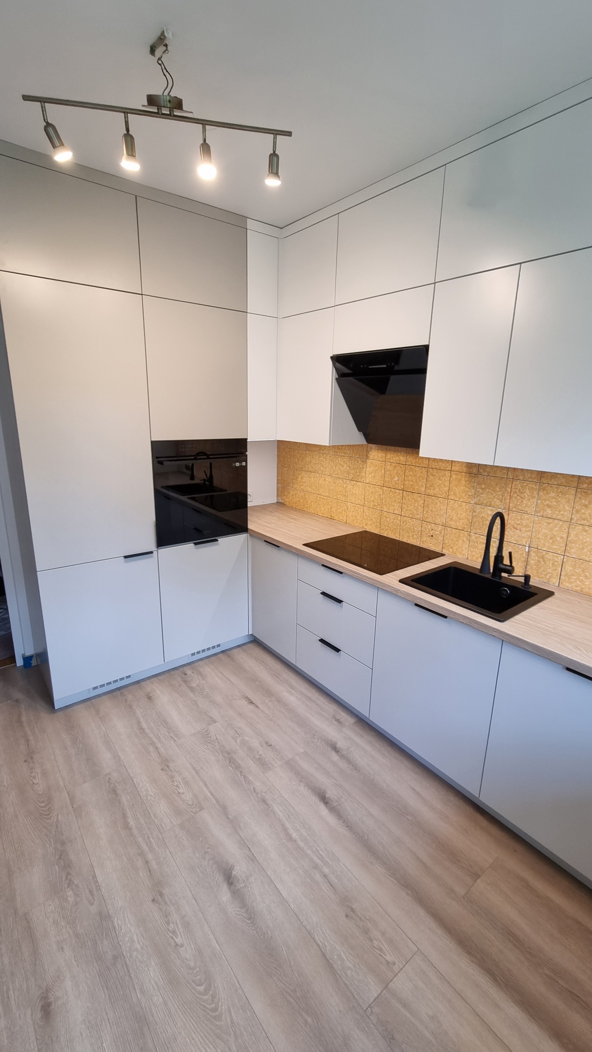 Nowoczesna, jasna kuchnia z białymi szafkami i drewnianym blatem, czarny zlew i płyta indukcyjna, oświetlenie punktowe.