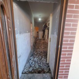 SolBud Usługi Ogólnobudowlane i Ziemne - Wnętrze w trakcie remontu: gruz na podłodze, ściany przygotowane do prac wykończeniowych, mężczyzna na drabinie z narzędziem w głębi korytarza.