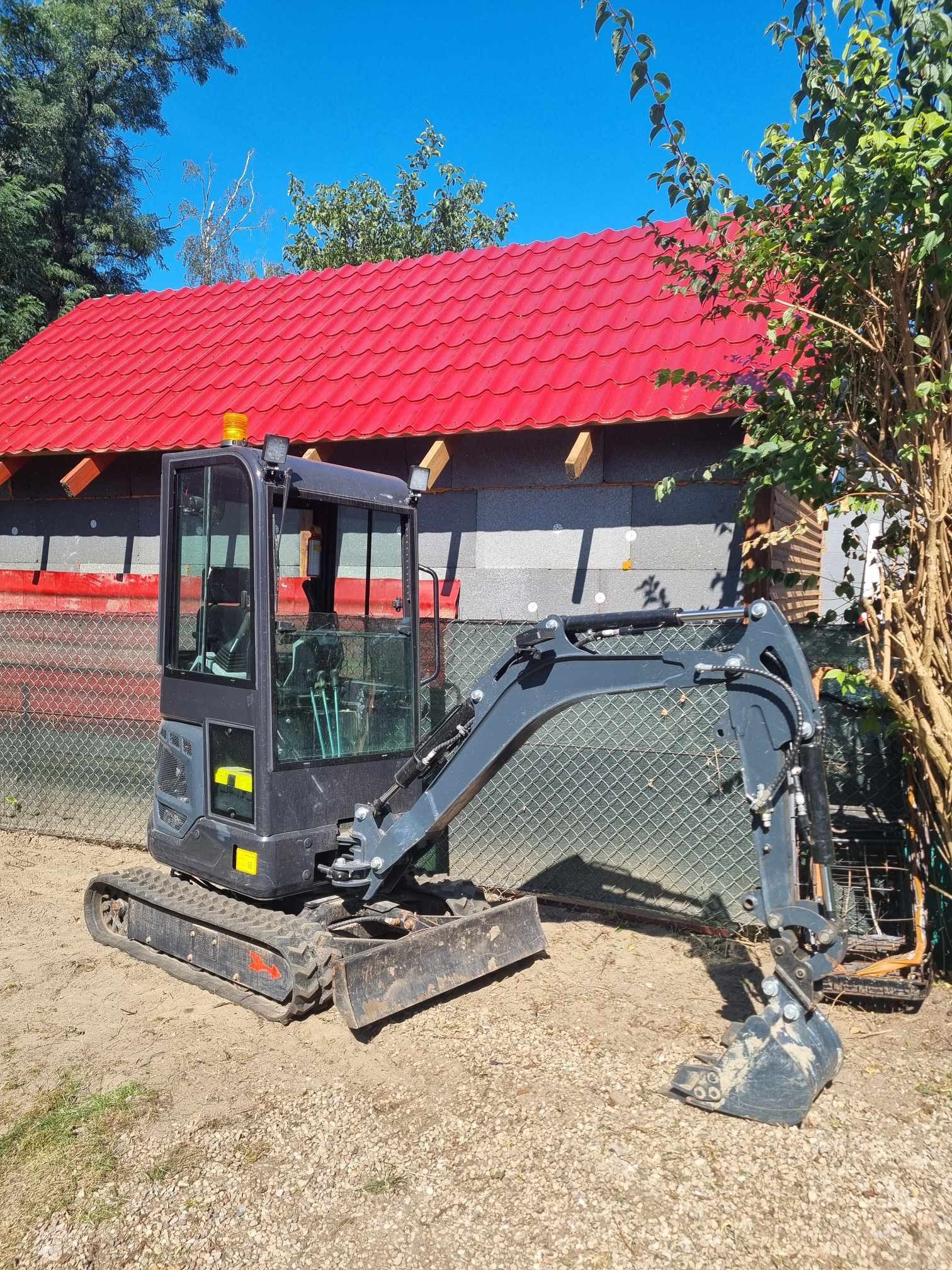 Mini koparka na terenie budowy w Silnowo, z widocznym, częściowo ocieplonym budynkiem z czerwoną blachodachówką w tle.
