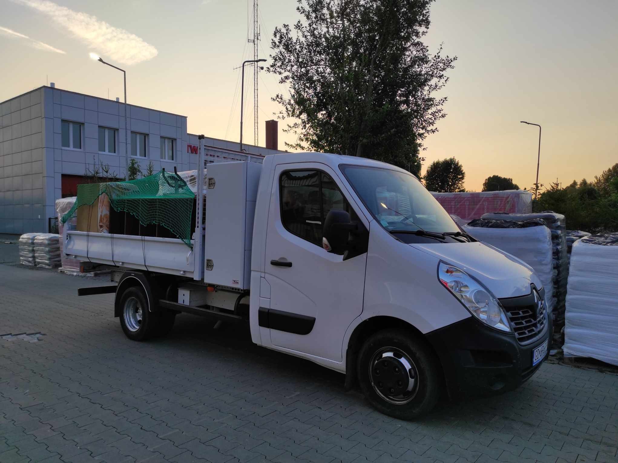 Biała ciężarówka typu pick-up z zieloną siatką zabezpieczającą ładunek, zaparkowana na brukowanym placu z widocznymi w tle workami z materiałami budowlanymi i budynkiem o zachodzie słońca.