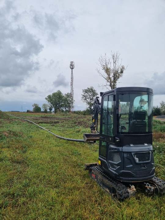 Mini koparka przy wykopie pod przewiert sterowany, widoczny rów i długa rura, w tle wieża telekomunikacyjna i pochmurne niebo.