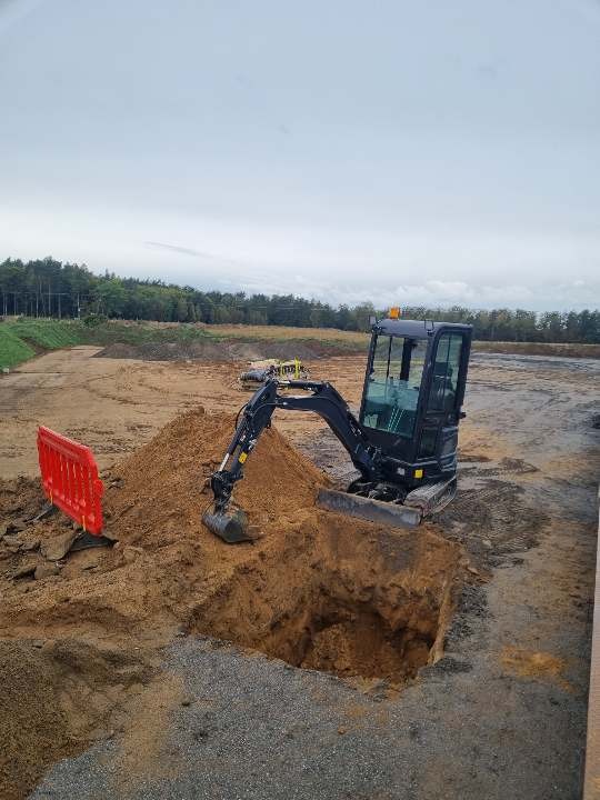 Mini koparka przy wykopie w piaszczystej ziemi, pomarańczowe barierki zabezpieczające, w tle zalesiony teren i pochmurne niebo.