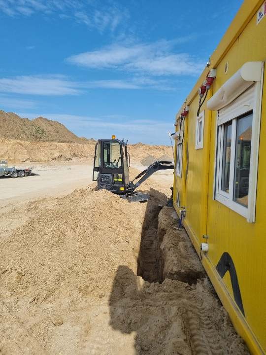 Mini koparka wykonuje wąski wykop w piaszczystym terenie obok żółtego kontenera budowlanego, widoczne ślady opon i hałda piasku, w tle wzgórza i błękitne niebo.