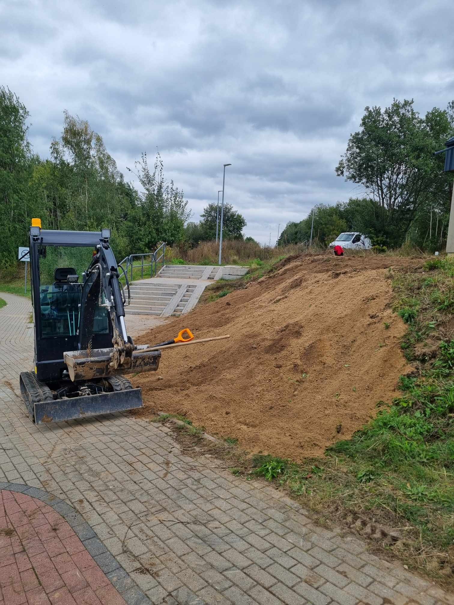 Czarny mini-koparka na kostce brukowej obok wzgórza piasku z widocznymi schodami i łopatą, w tle zaparkowany biały samochód dostawczy.
