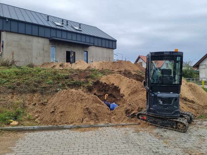 Mała czarna koparka stoi obok wykopu w ziemi, w którym widać pracownika z łopatą, w tle budynek w stanie surowym.