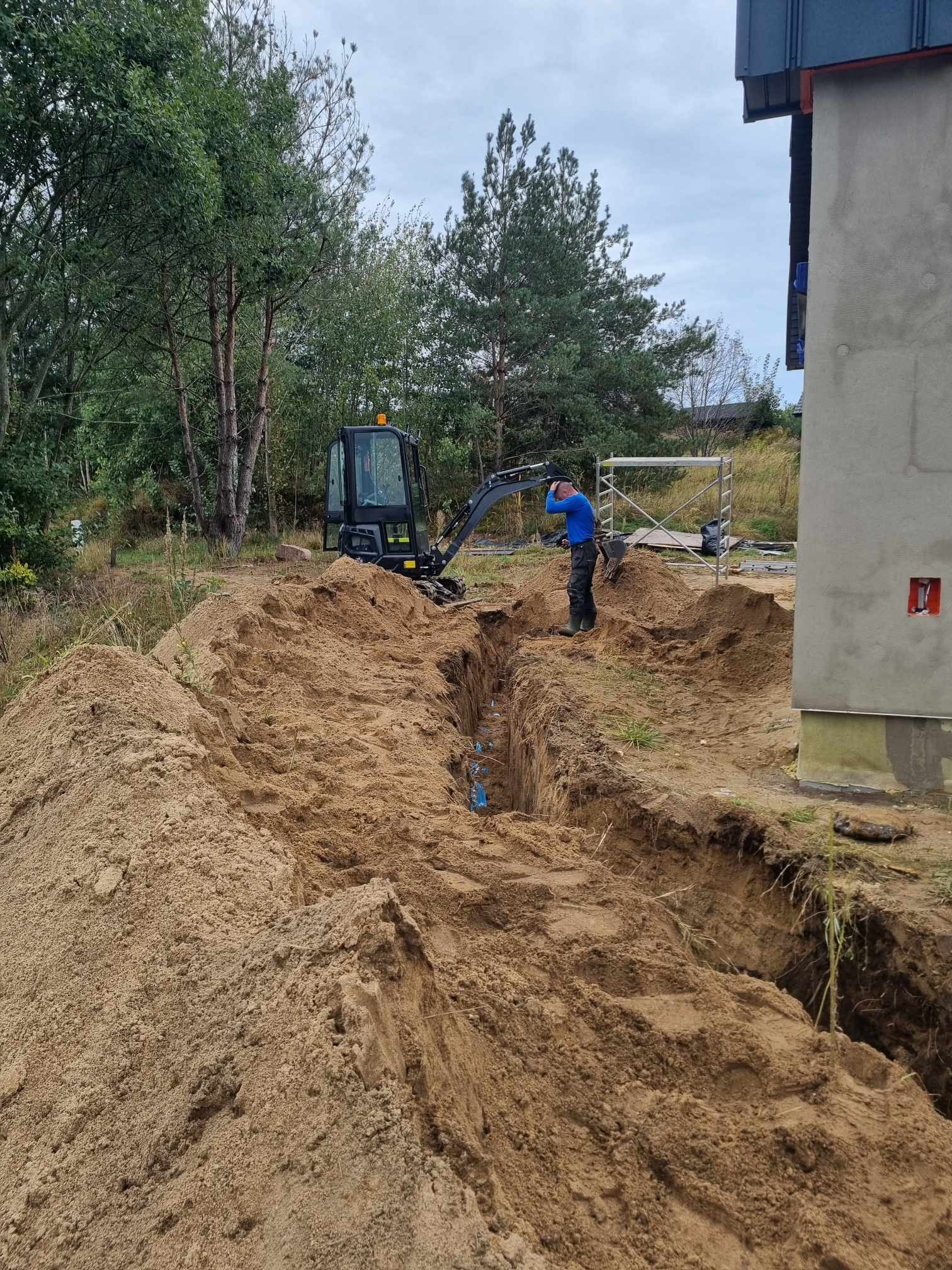 Wąski wykop w piaszczystej ziemi z ułożonymi niebieskimi rurami, minikoparka w tle, pracownik w niebieskiej koszulce przygląda się pracy, obok ściana budynku w trakcie budowy i rusztowanie.