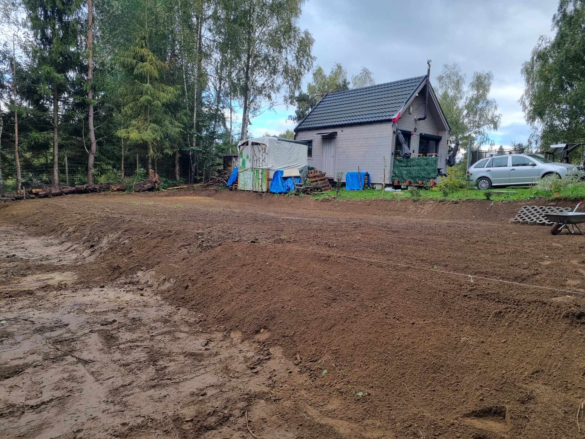 Przygotowany teren pod budowę, wyrównana ziemia z widocznymi śladami opon, w tle drewniany domek i samochód osobowy.