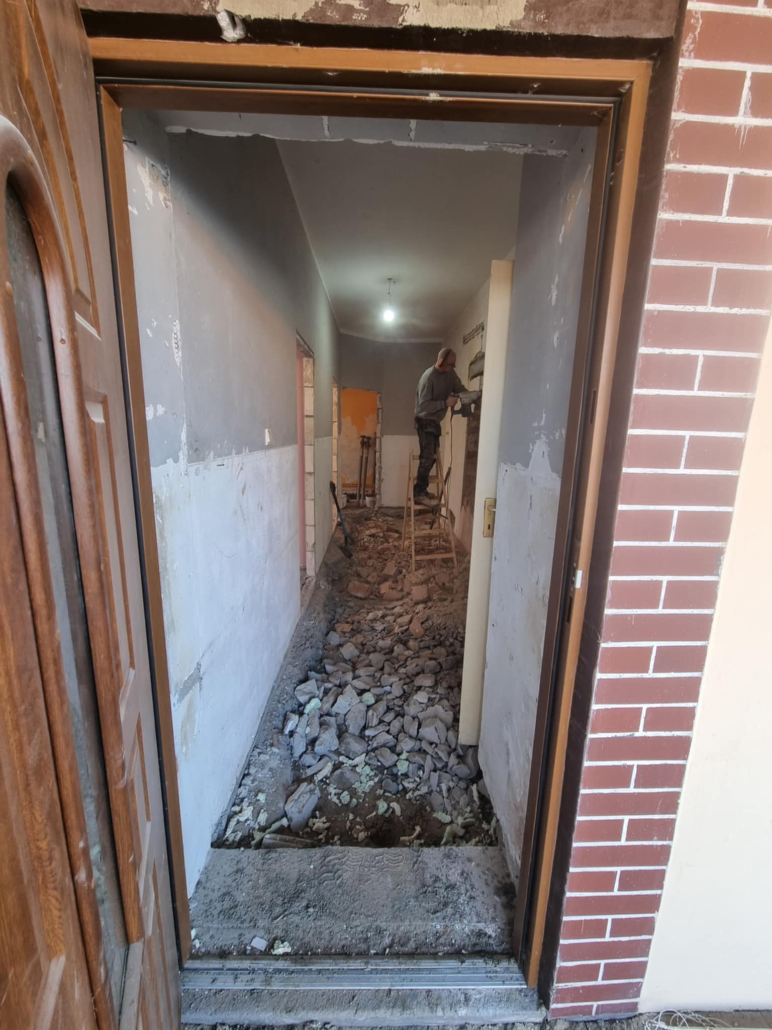 Wnętrze w trakcie remontu: gruz na podłodze, ściany przygotowane do prac wykończeniowych, mężczyzna na drabinie z narzędziem w głębi korytarza.