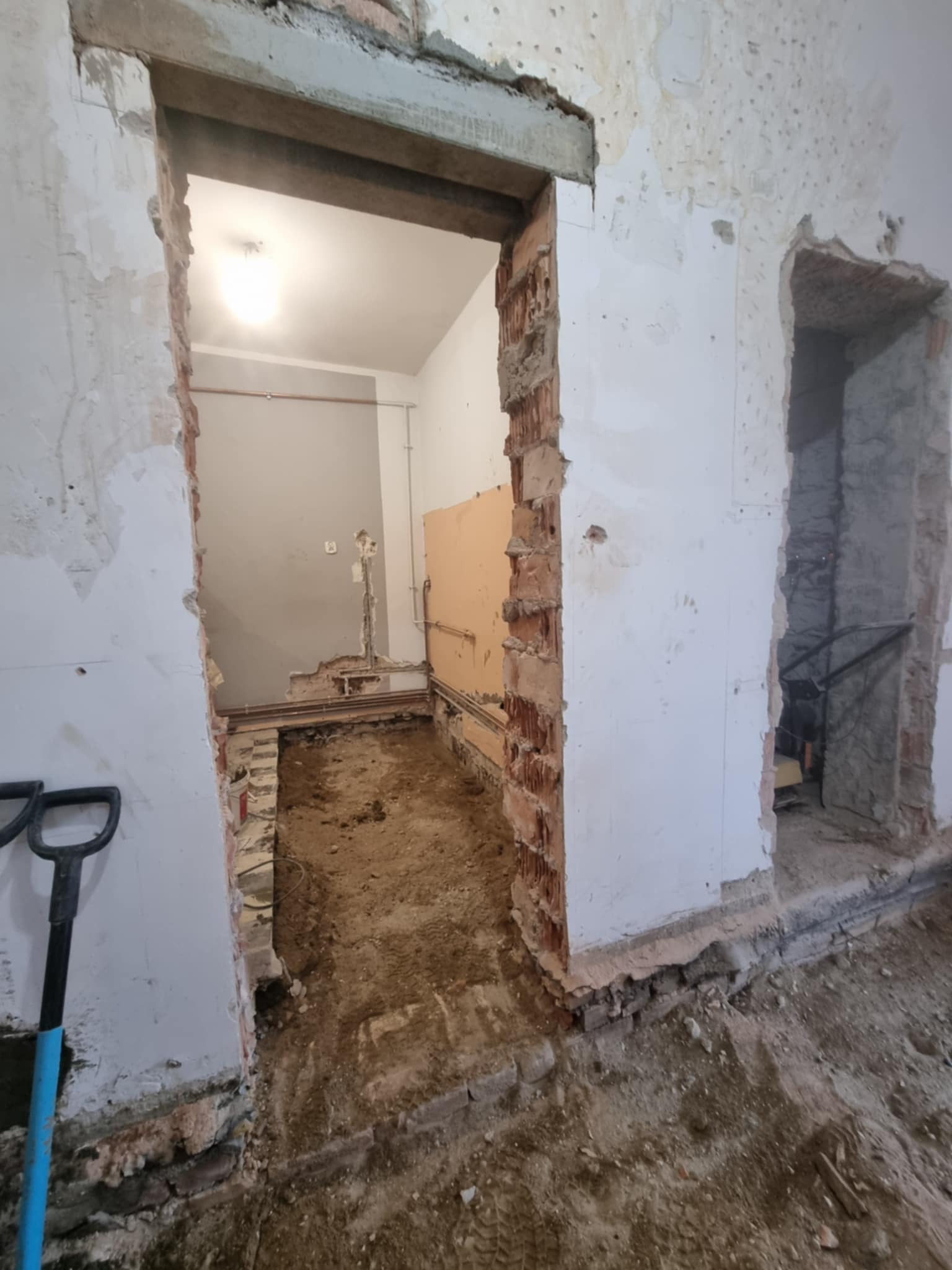 Wnętrze w trakcie remontu: odsłonięte cegły, instalacje i ziemia na podłodze. Widoczne ślady po kuciu ścian i przygotowania do dalszych prac wykończeniowych.