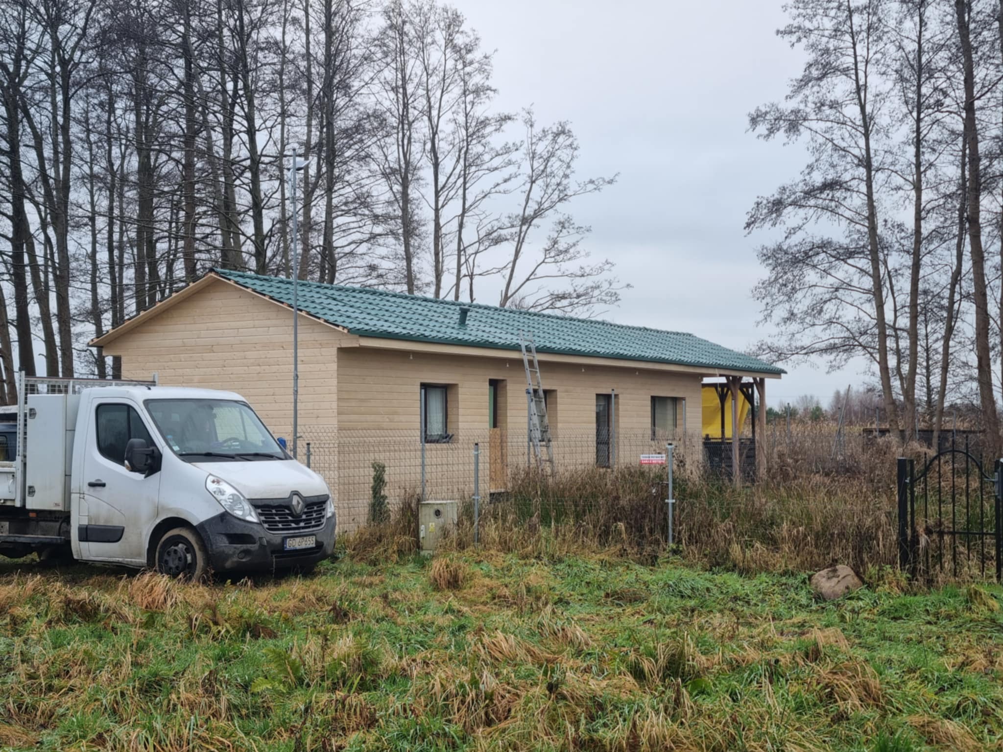 Nowo wybudowany, parterowy dom z zielonym dachem, drabina oparta o ścianę, biały samochód dostawczy na pierwszym planie, jesienne otoczenie.