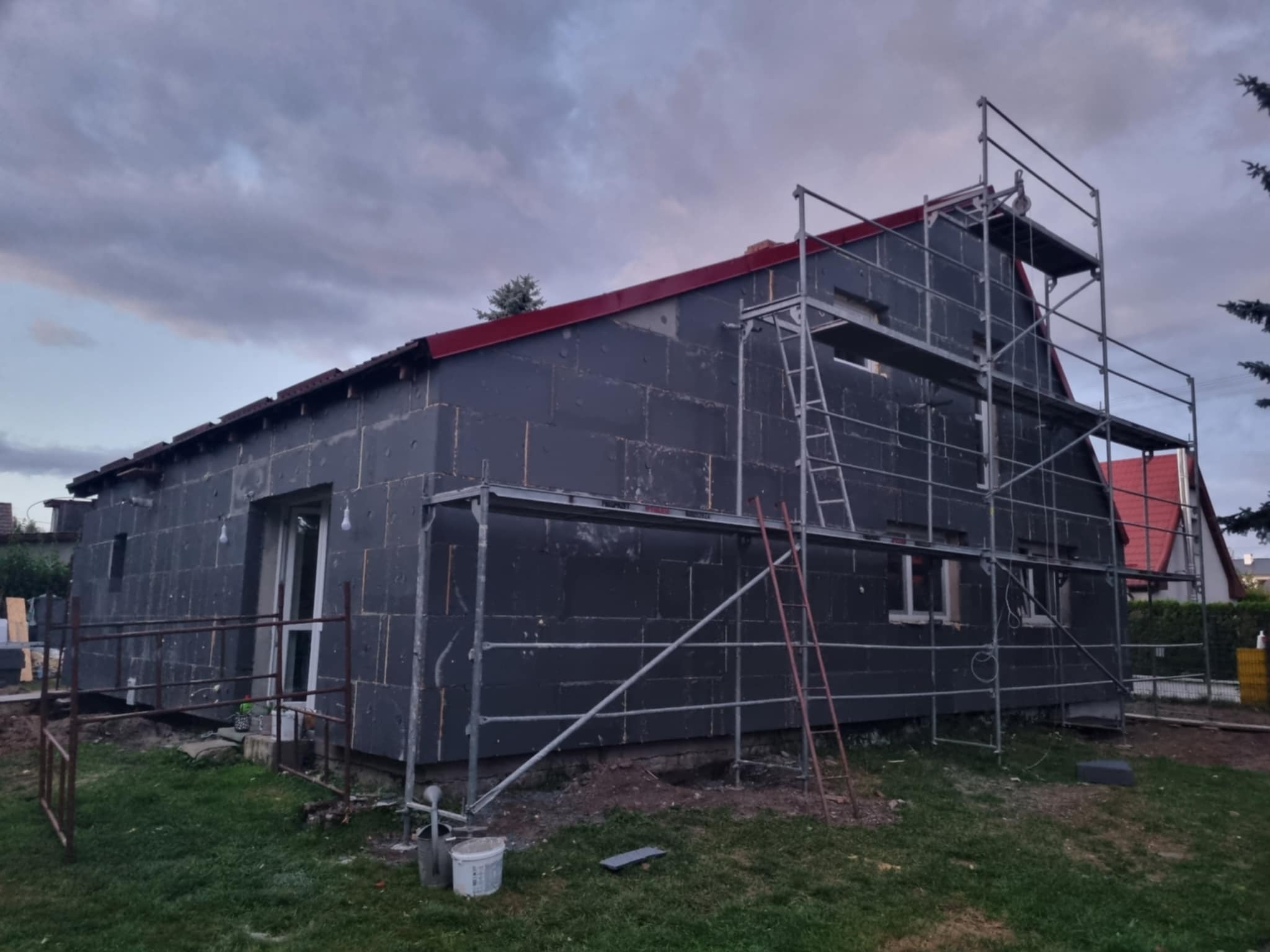 Dom w trakcie ocieplania styropianem grafitowym, widoczne rusztowania i fragment dachu z czerwoną blachą. Prace budowlane w toku, widok z zewnątrz na elewację.