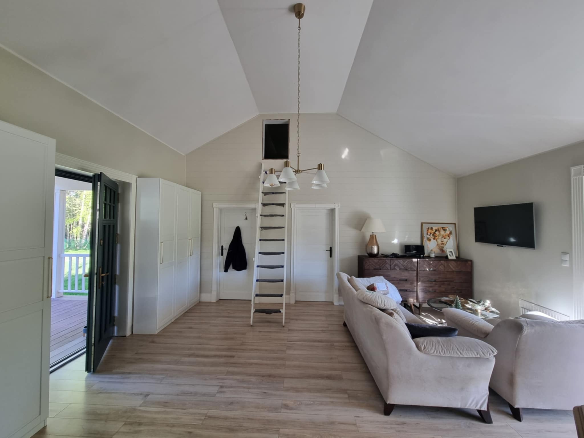 Jasne wnętrze z drewnianą podłogą, białymi szafami i schodami prowadzącymi do okna dachowego. Widoczny salon z sofami i komodą.