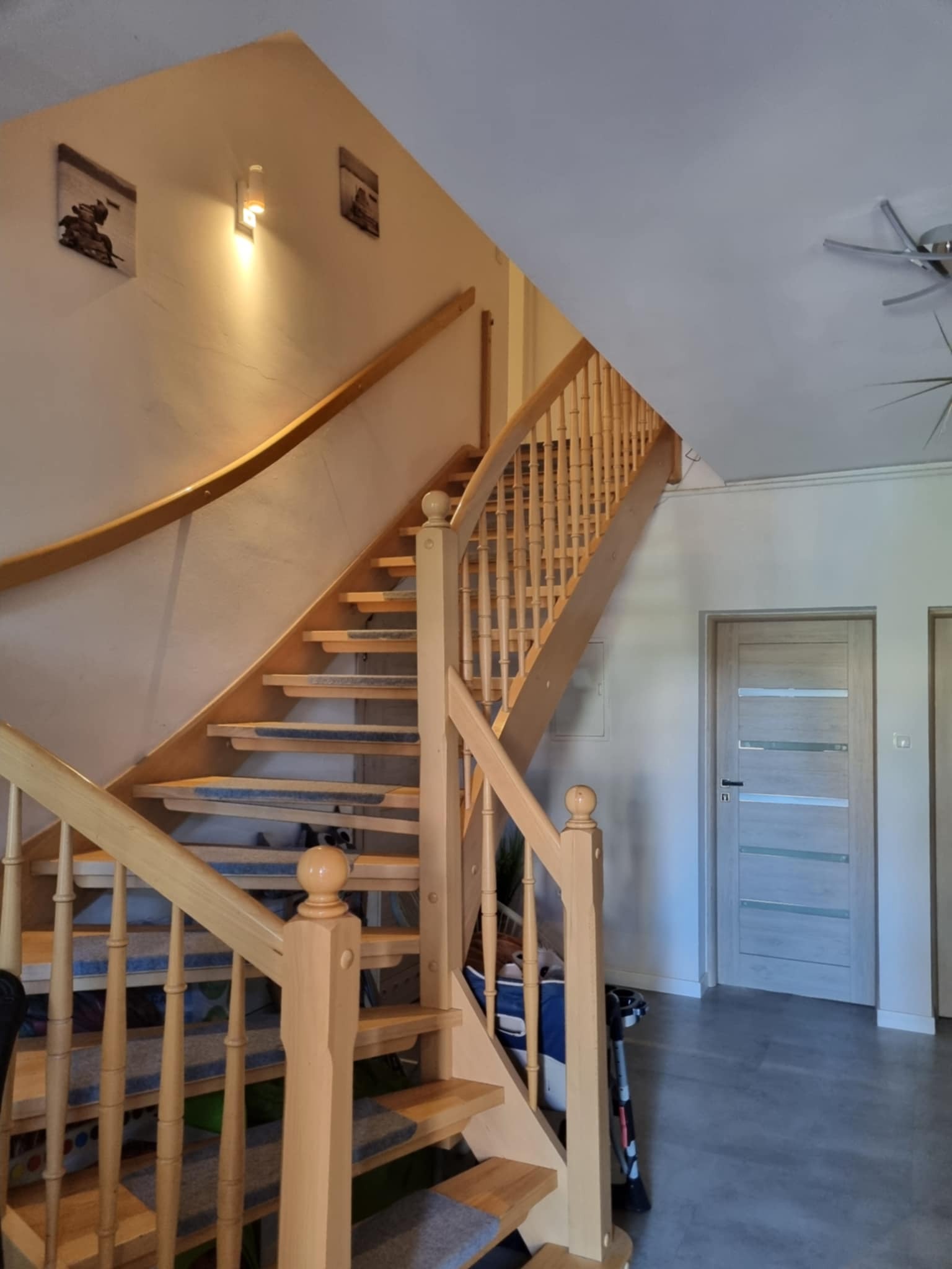Jasne, drewniane schody z dywanikami na stopniach, prowadzące na piętro w domu. Widoczne balustrady i fragment drzwi. Ciepłe oświetlenie.