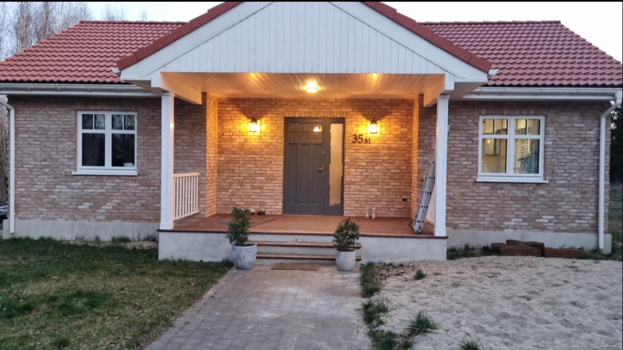 Elewacja domu z cegły klinkierowej, oświetlony ganek z numerem 35M, widok z brukowanego chodnika na trawnik z piaskiem. Dom po remoncie.