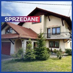 Nikodem Ankudowicz REMAX INVEST - Dwupiętrowy dom z czerwonym dachem i balkonem z kutą balustradą, oznaczony dużym czerwonym napisem 'SPRZEDANE' na niebieskim tle.