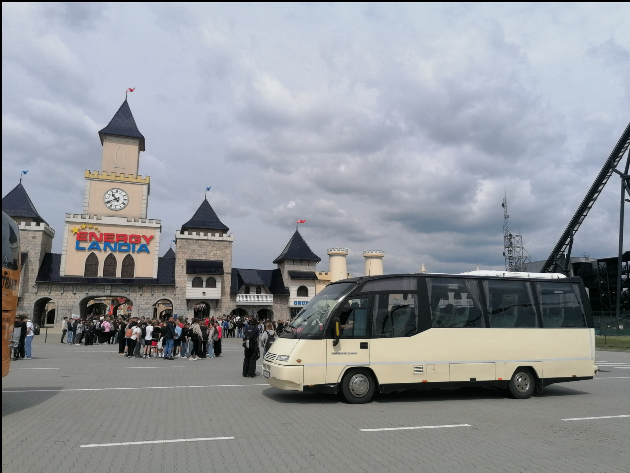 Beżowy bus stoi przed wejściem do Energylandii, obok grupka ludzi. W tle wieże zamku z zegarem i element kolejki górskiej. Pochmurne niebo.