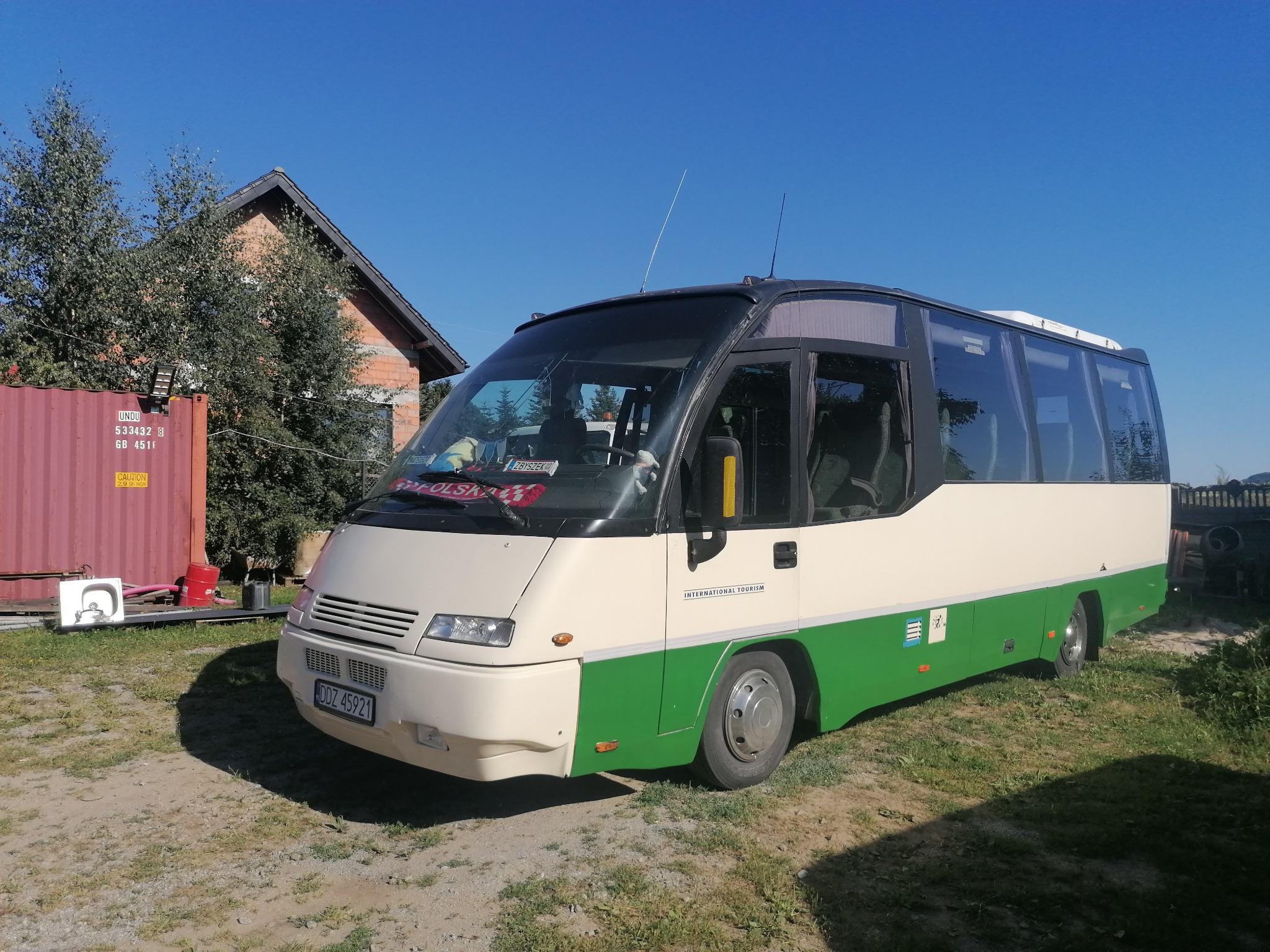 Autobus turystyczny z napisem 'International Tourism' na tle błękitnego nieba i zieleni. Kontener i budynek w tle. Polski akcent na przedniej szybie, antena na dachu.