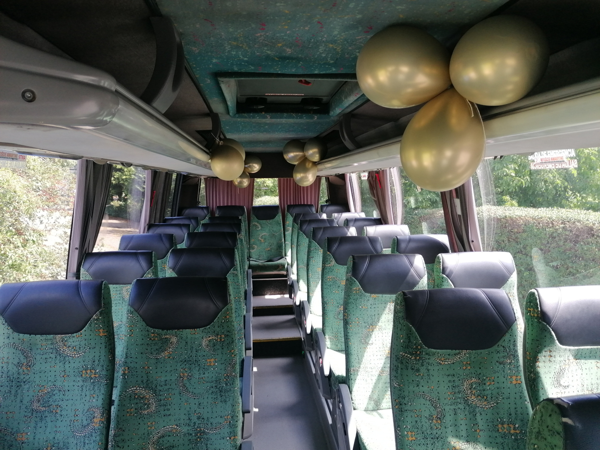 Wnętrze autobusu z zielonymi fotelami, udekorowane złotymi balonami, sugerujące wynajem na imprezę okolicznościową. Widok na rząd foteli i okna.