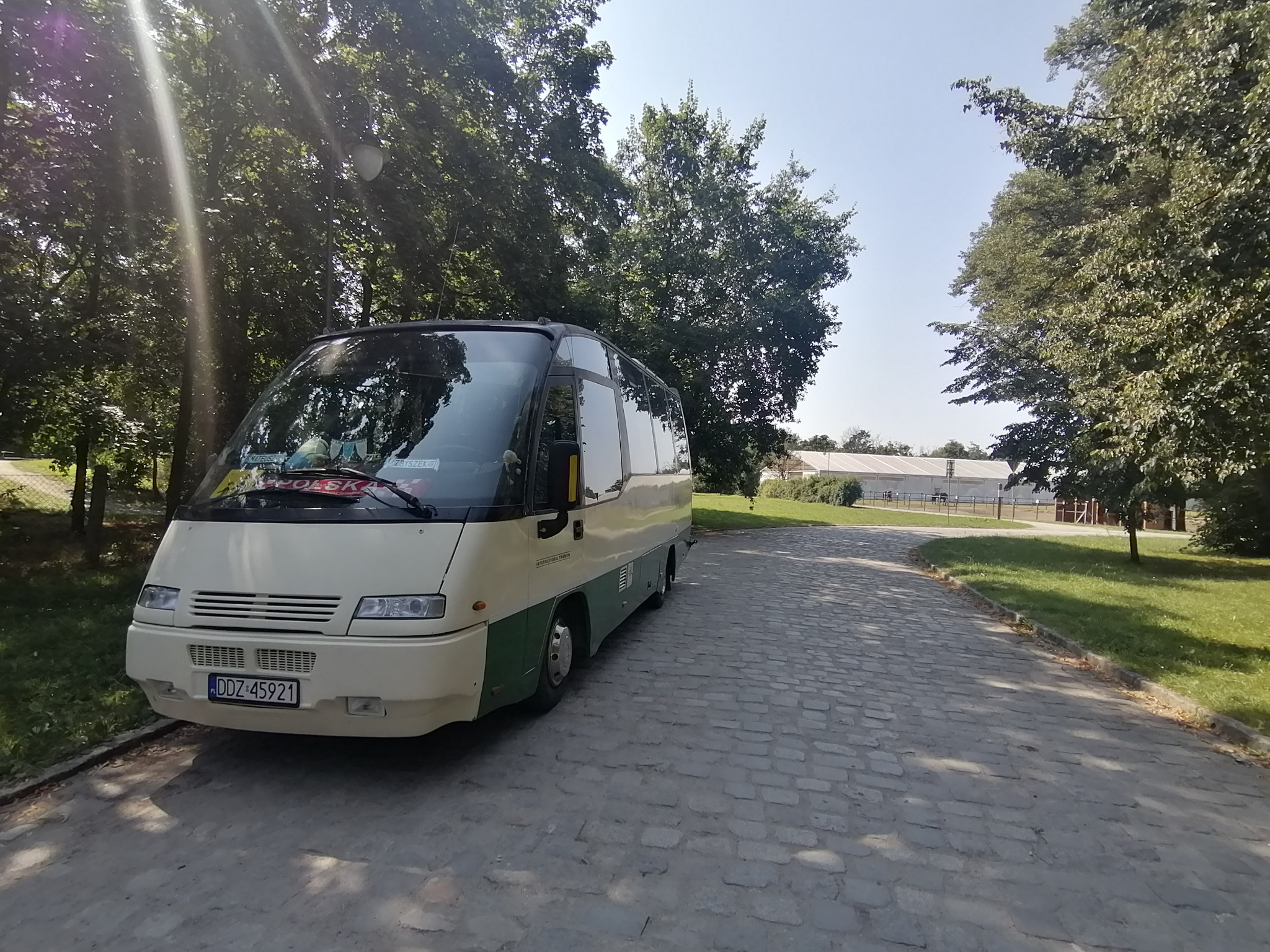 Beżowo-zielony bus z napisem 'Polska' za szybą, zaparkowany na brukowanej drodze w otoczeniu drzew. Widoczna tablica rejestracyjna DDZ 45921 i budynek w tle.