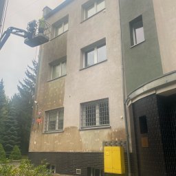 SPECTROBUD sp. z o.o. - Pracownik na podnośniku koszowym myje elewację budynku, widoczna różnica między umytą a brudną częścią ściany, żółta skrzynka gazowa przy budynku.