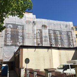 SPECTROBUD sp. z o.o. - Budynek w trakcie prac elewacyjnych, osłonięty siatką z reklamami firm Ceresit i Caparol, widoczny fragment rusztowania i przenośna toaleta.