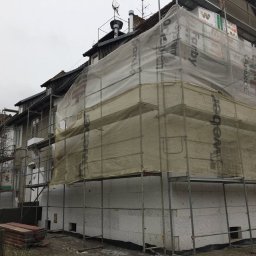 SPECTROBUD sp. z o.o. - Budynek wielorodzinny w trakcie ocieplania elewacji styropianem, zabezpieczony siatką i rusztowaniem, widoczne materiały budowlane Weber i Farby System.