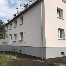 SPECTROBUD sp. z o.o. - Odnowiony budynek wielorodzinny z jasną elewacją i szarą podmurówką, widoczne rury spustowe i nowa kostka brukowa wokół budynku.