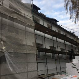 SPECTROBUD sp. z o.o. - Budynek w trakcie prac elewacyjnych, widoczne rusztowania, siatka ochronna i materiały izolacyjne, pracownik przy pracy na rusztowaniu.