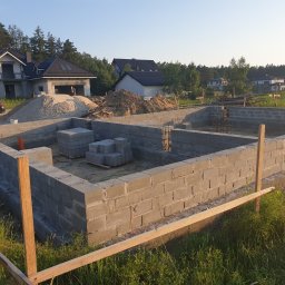 SPECTROBUD sp. z o.o. - Fundamenty z bloczków betonowych w trakcie budowy domu jednorodzinnego, widoczne zbrojenia i drewniane szalunki, w tle częściowo wykończony dom z czarnym dachem i sterty piasku.