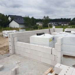 SPECTROBUD sp. z o.o. - Wznoszenie ścian parteru domu z białych bloczków silikatowych; widoczne palety z zapakowanymi bloczkami, fragmenty ścian oraz otoczenie z innymi budynkami i zielenią w tle.