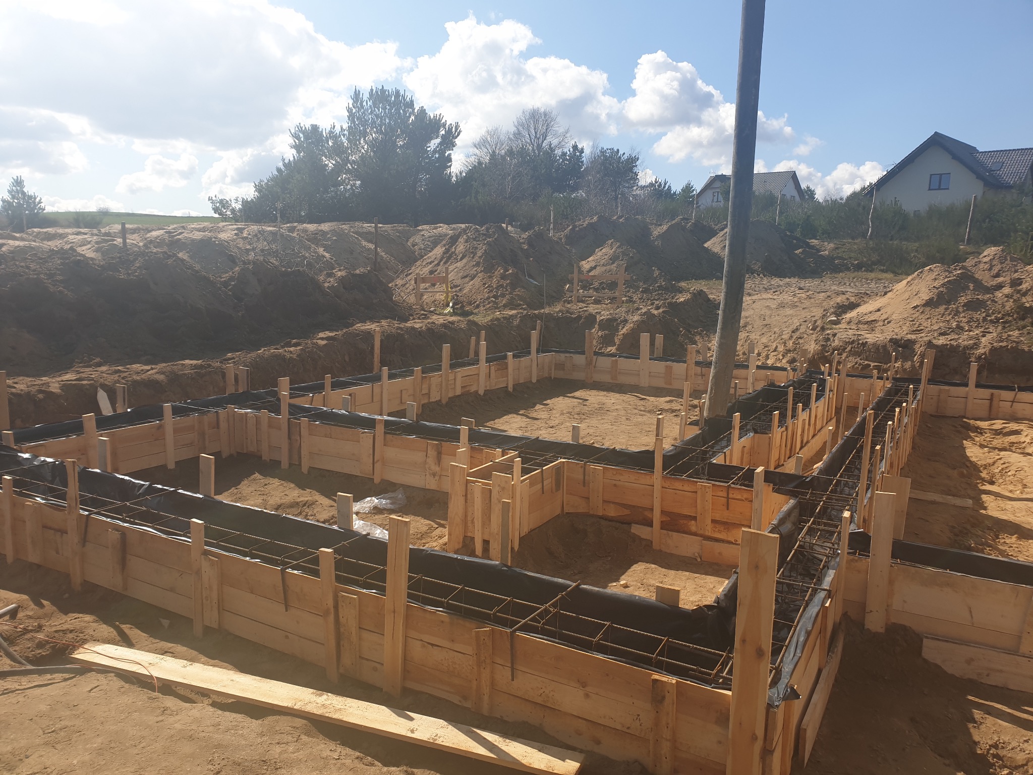 Przygotowanie szalunku i zbrojenia pod fundament domu murowanego, widoczne drewniane deski, czarna folia i stalowe pręty zbrojeniowe, w tle wykopana ziemia i dom jednorodzinny pod błękitnym niebem.