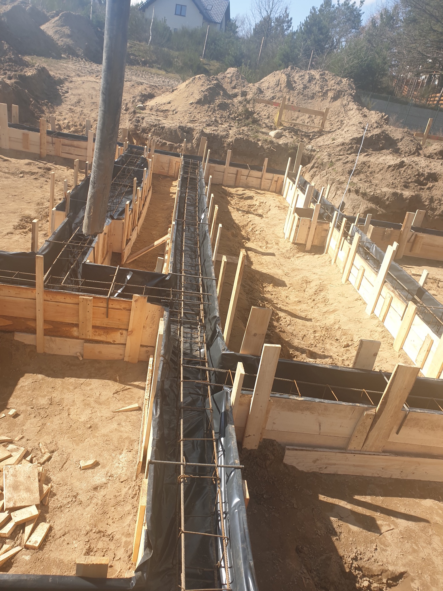 Wylewanie betonu do szalunków zbrojeniowych fundamentów domu jednorodzinnego na piaszczystym terenie.