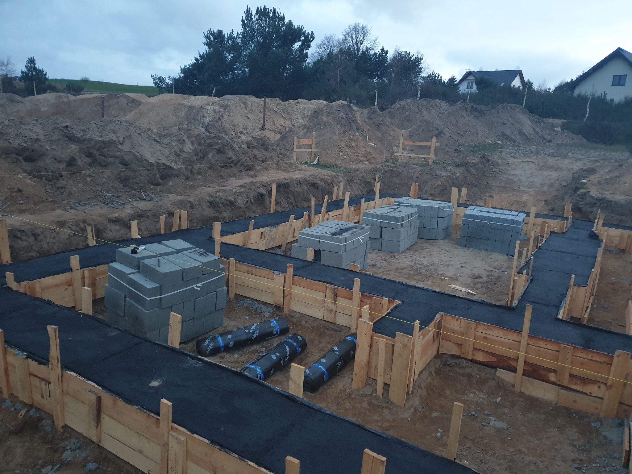 Fundamenty domu w trakcie budowy: drewniane szalunki, izolacja przeciwwilgociowa i bloczki betonowe na działce z widokiem na domy w tle.