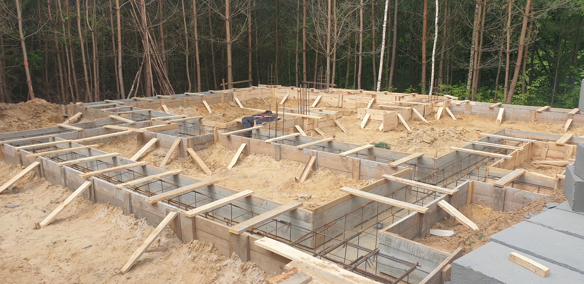 Przygotowanie fundamentów pod dom murowany: drewniane szalunki zbrojone stalą na piaszczystym podłożu, w tle widoczny las.