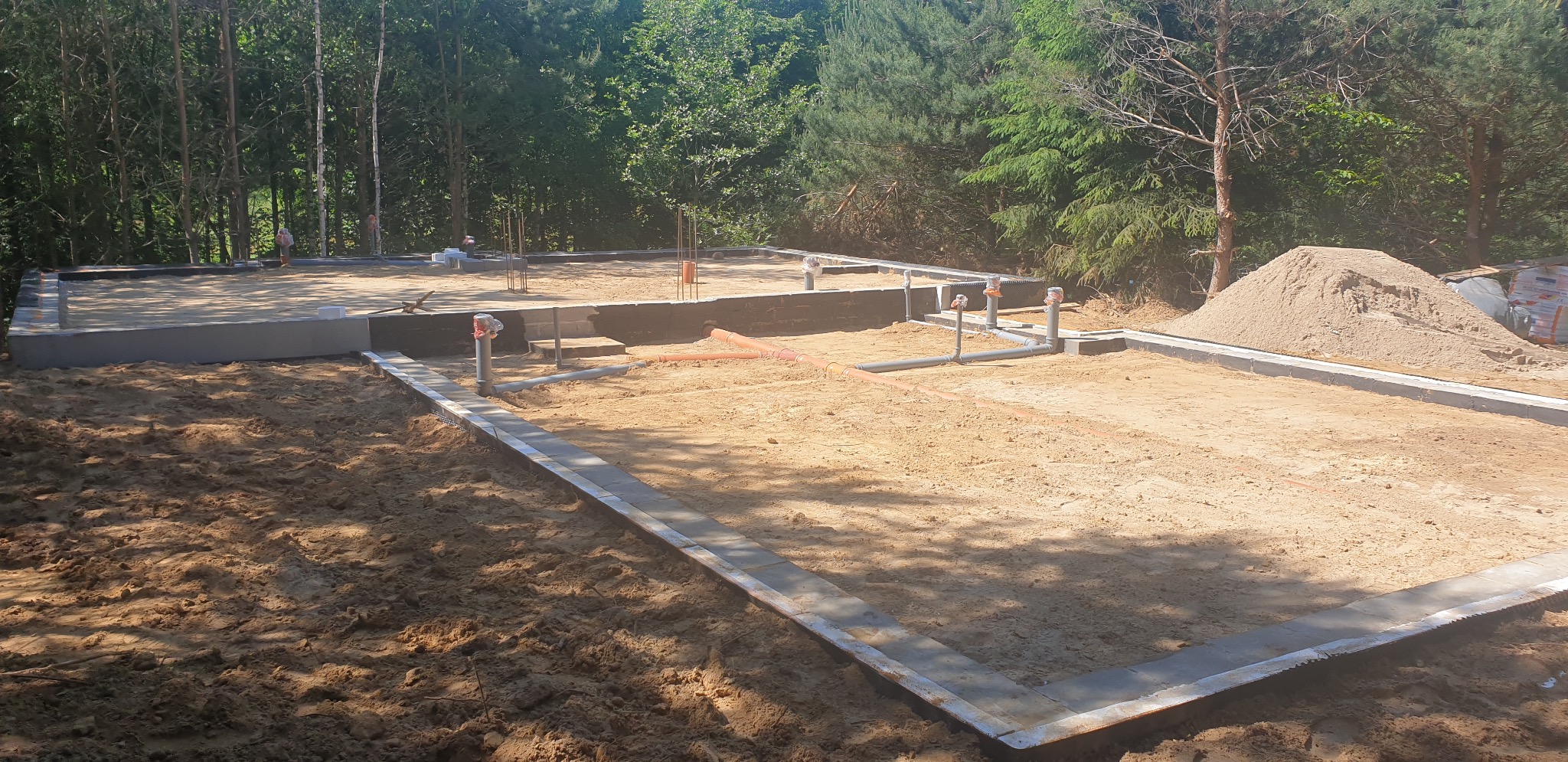 Fundamenty domu w trakcie budowy, widoczne zbrojenia, izolacja pionowa i instalacje kanalizacyjne, w tle las.