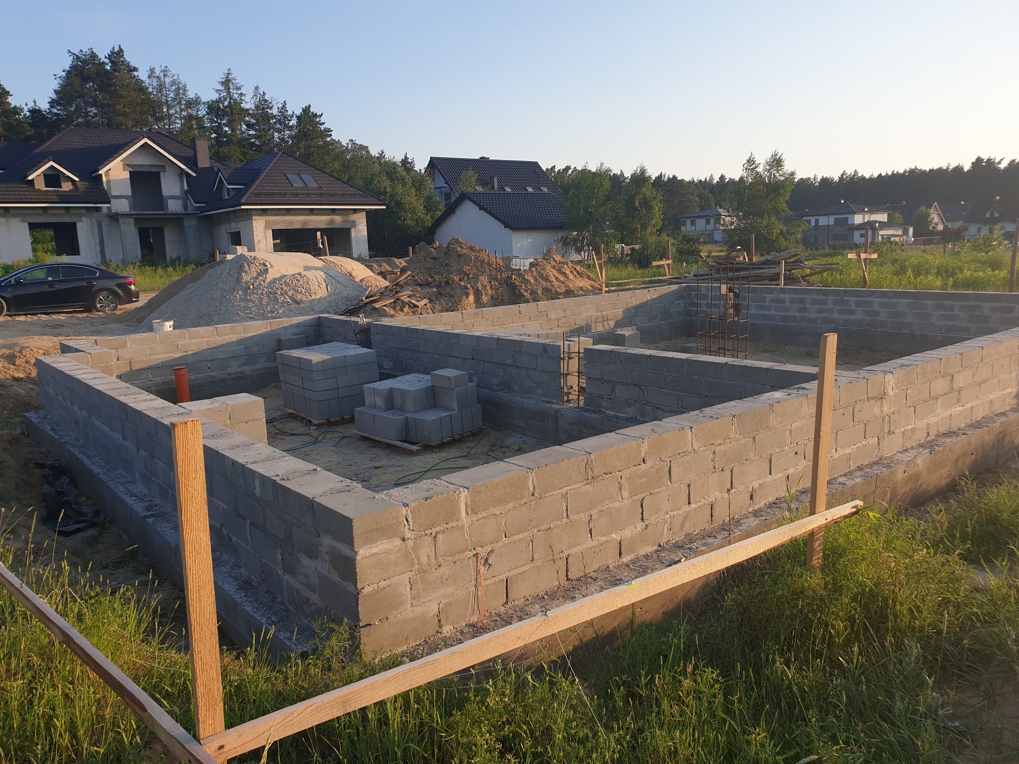 Fundamenty z bloczków betonowych w trakcie budowy domu jednorodzinnego, widoczne zbrojenia i drewniane szalunki, w tle częściowo wykończony dom z czarnym dachem i sterty piasku.