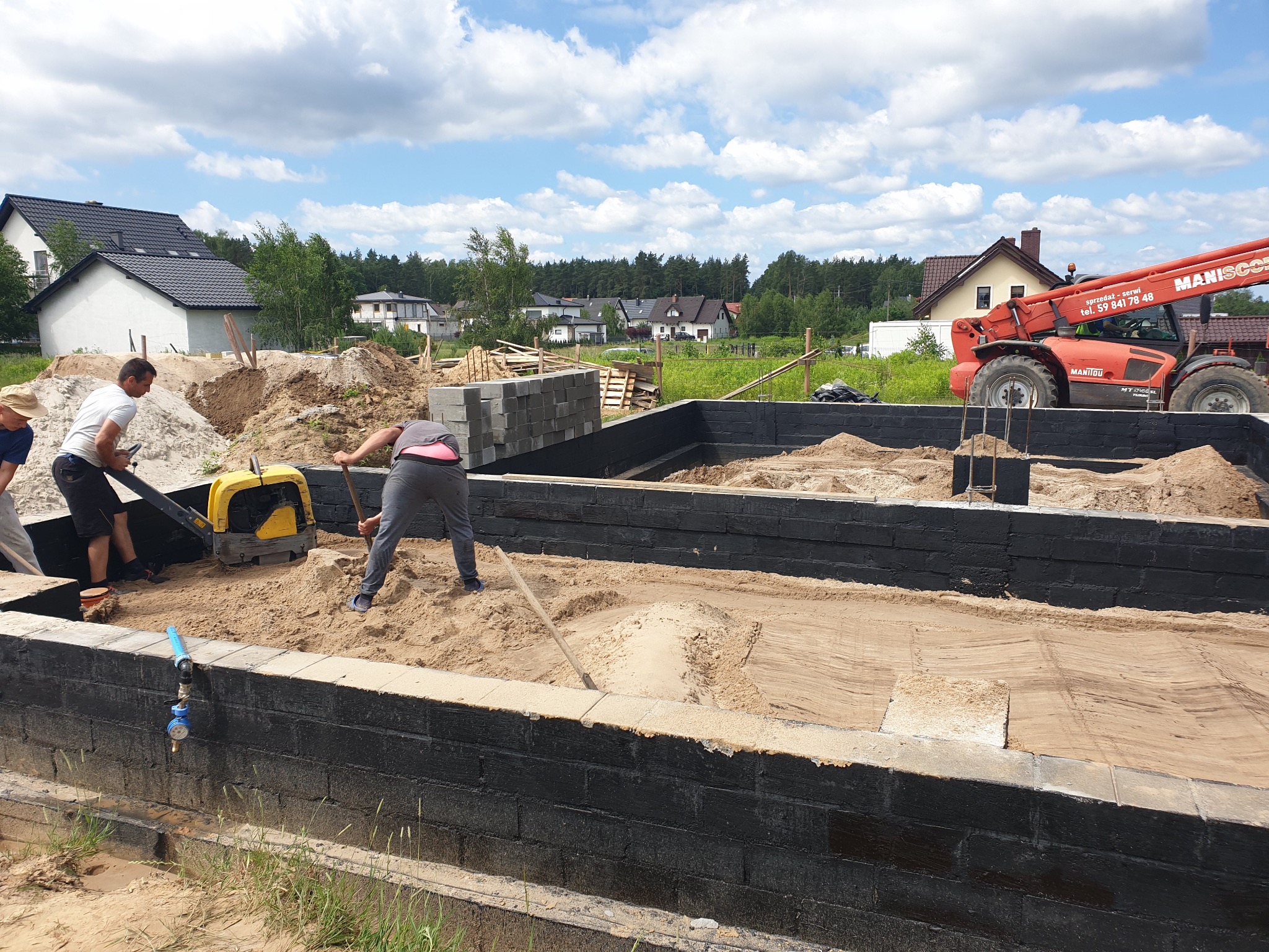 Fundamenty domu z izolacją, przygotowywane pod wylewkę, piasek ubijany zagęszczarką i wyrównywany łopatą, w tle podnośnik teleskopowy Manitou.