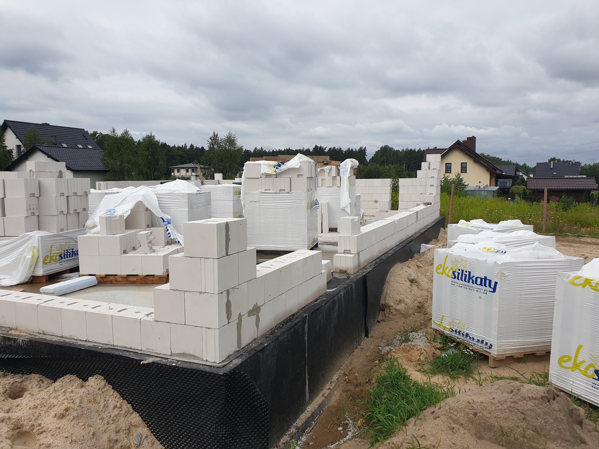 Budowa domu z białych bloczków silikatowych, widoczne fundamenty izolowane czarną folią, palety z bloczkami Eko Silikaty w tle zabudowa jednorodzinna.