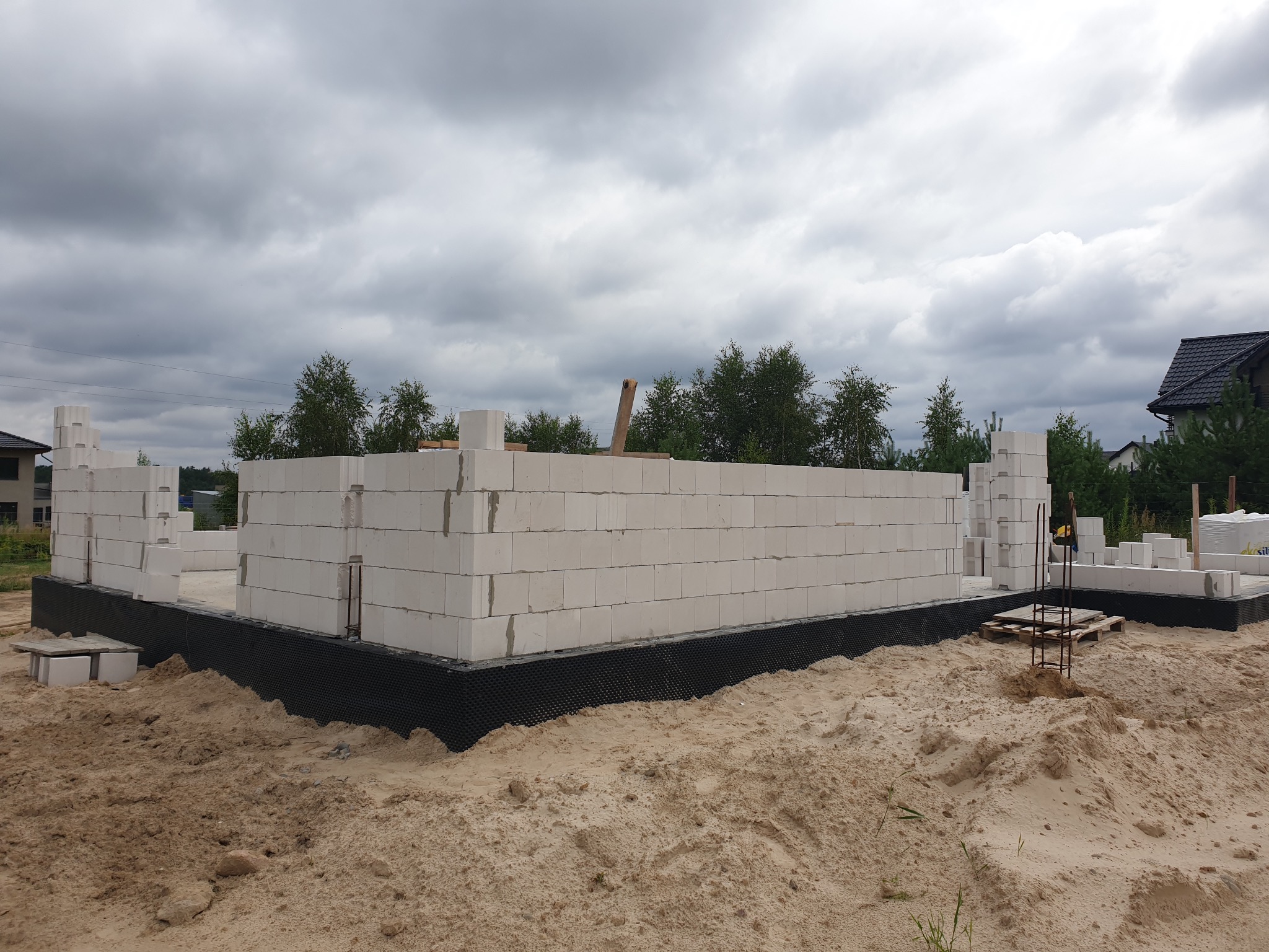 Wznoszenie ścian parteru domu z białych bloczków betonu komórkowego Ytong Silka, zabezpieczonych czarną folią hydroizolacyjną na fundamencie, w tle zachmurzone niebo i drzewa.