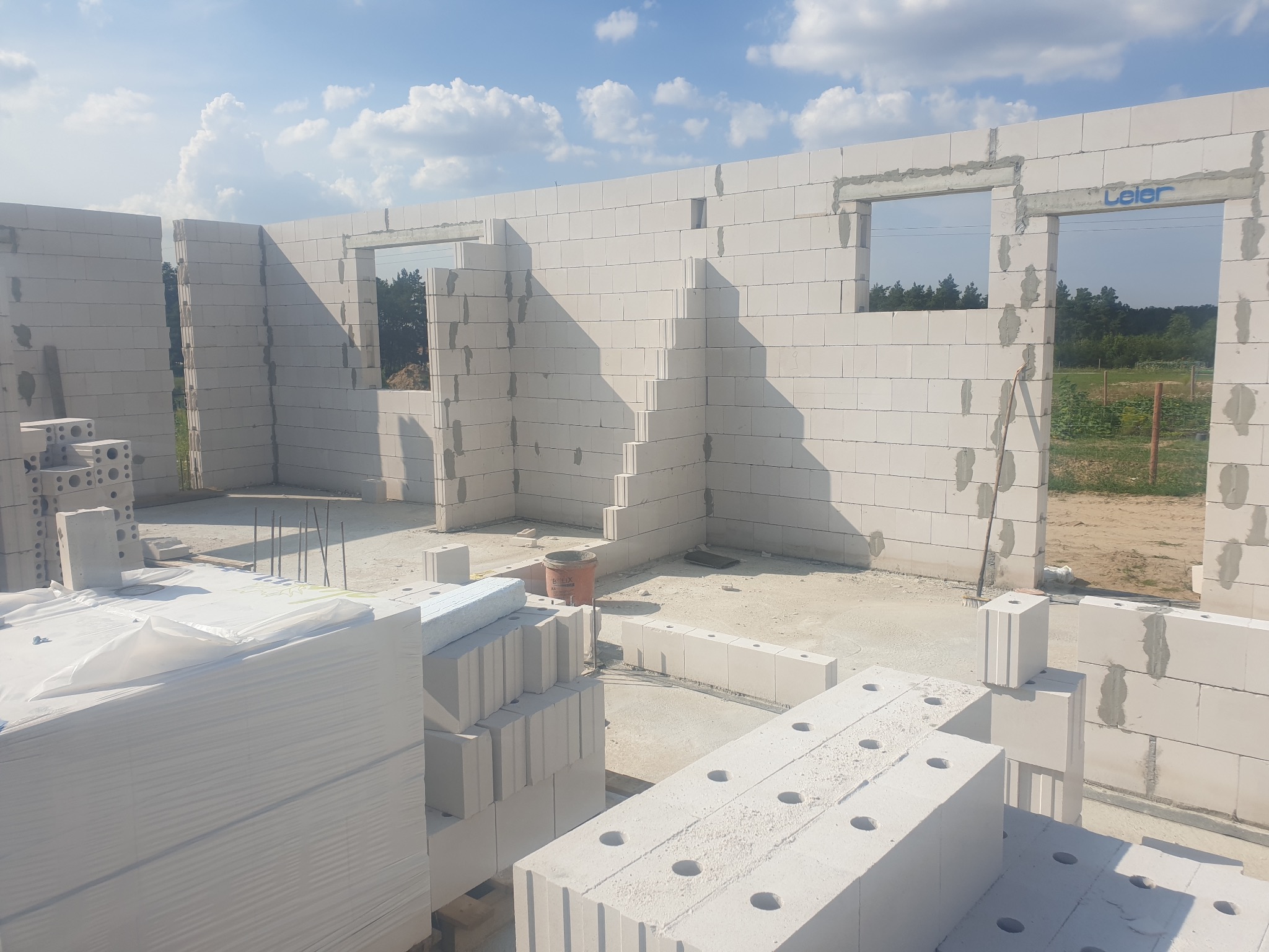 Wznoszenie ścian parterowego domu z białych bloczków, widoczne otwory okienne i drzwiowe, w tle zieleń drzew.