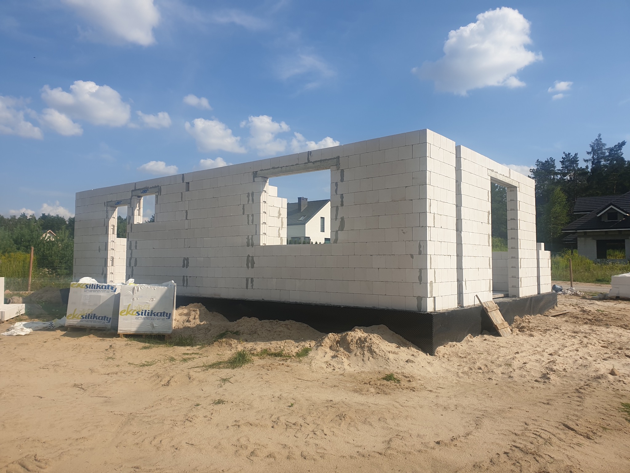 Wznoszenie ścian parterowego domu z białych bloczków silikatowych; widoczne okna i drzwi, zabezpieczenie fundamentów czarną folią oraz palety z materiałem budowlanym.