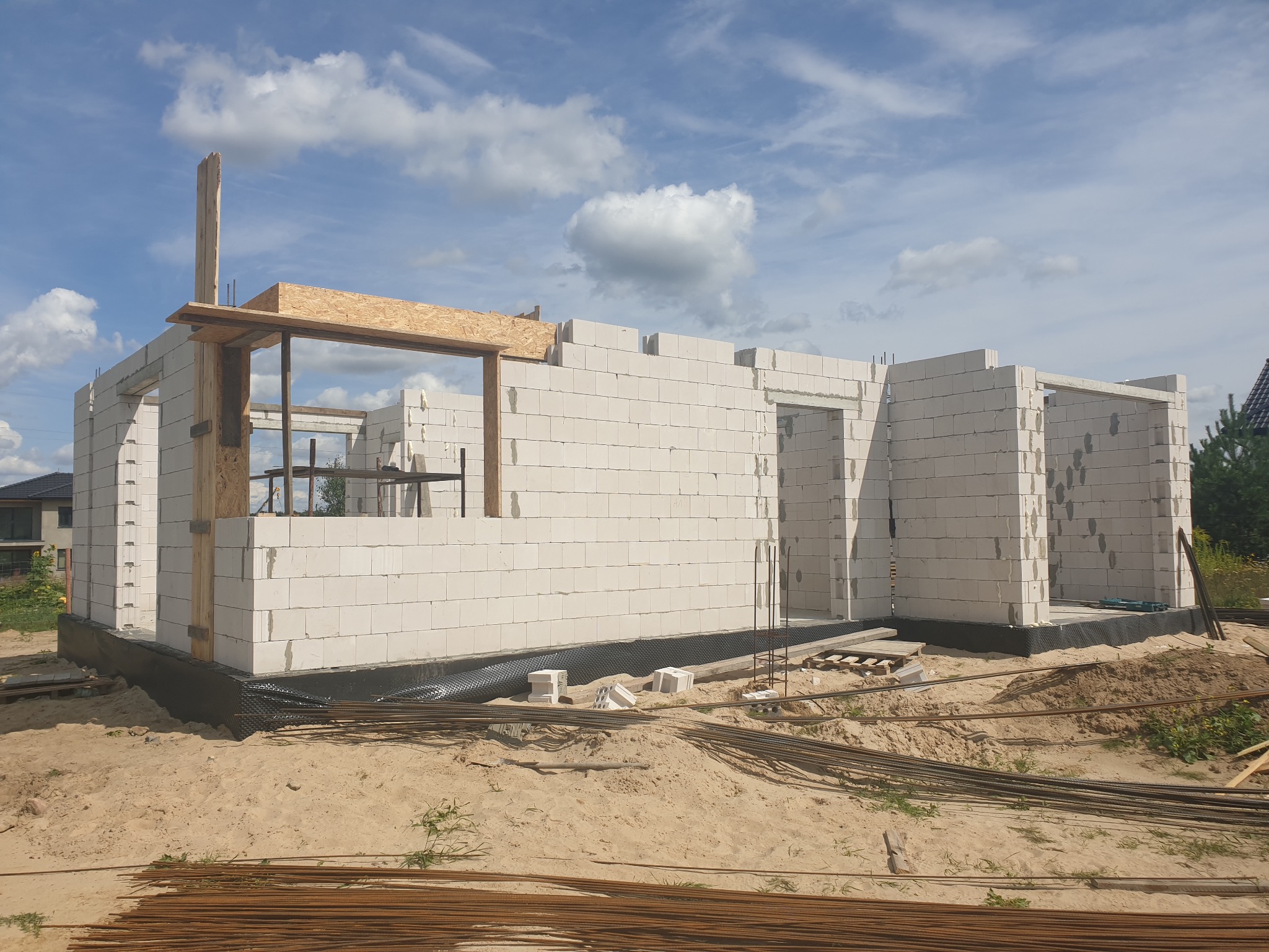 Wznoszenie ścian parterowego domu z białych bloczków, widoczne zbrojenia i drewniane elementy szalunku, teren budowy w stanie surowym otwartym.