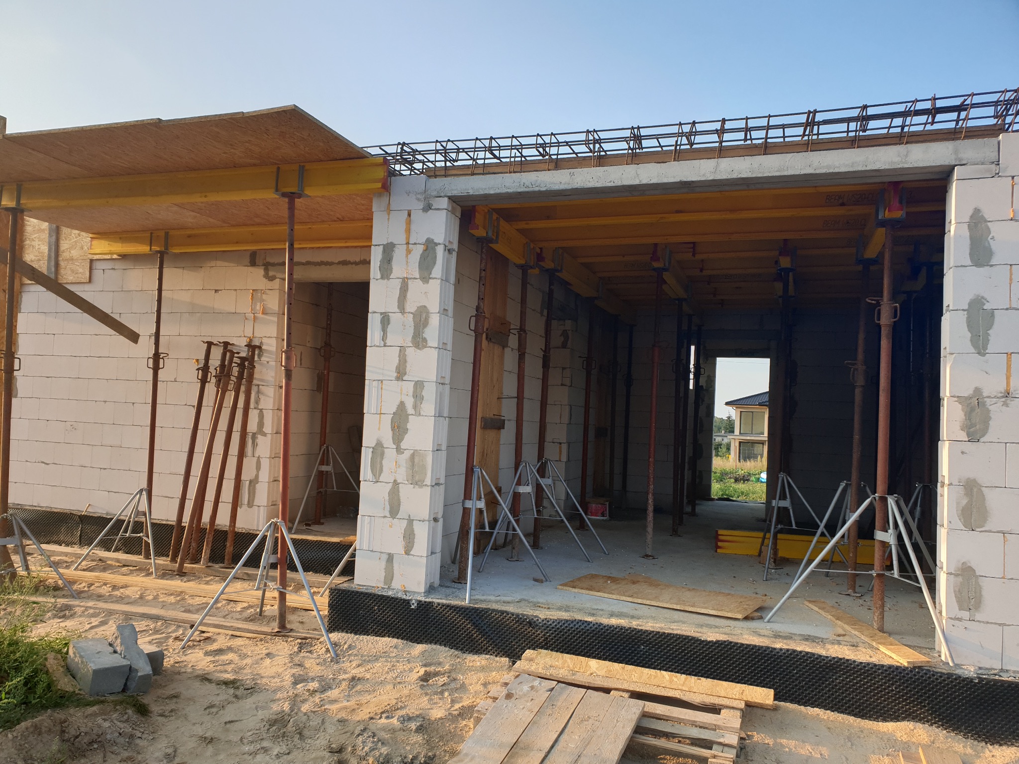 Wznoszenie ścian parterowego domu z betonu komórkowego, widoczne podpory stropowe, zbrojenie wieńca i czarna folia fundamentowa.