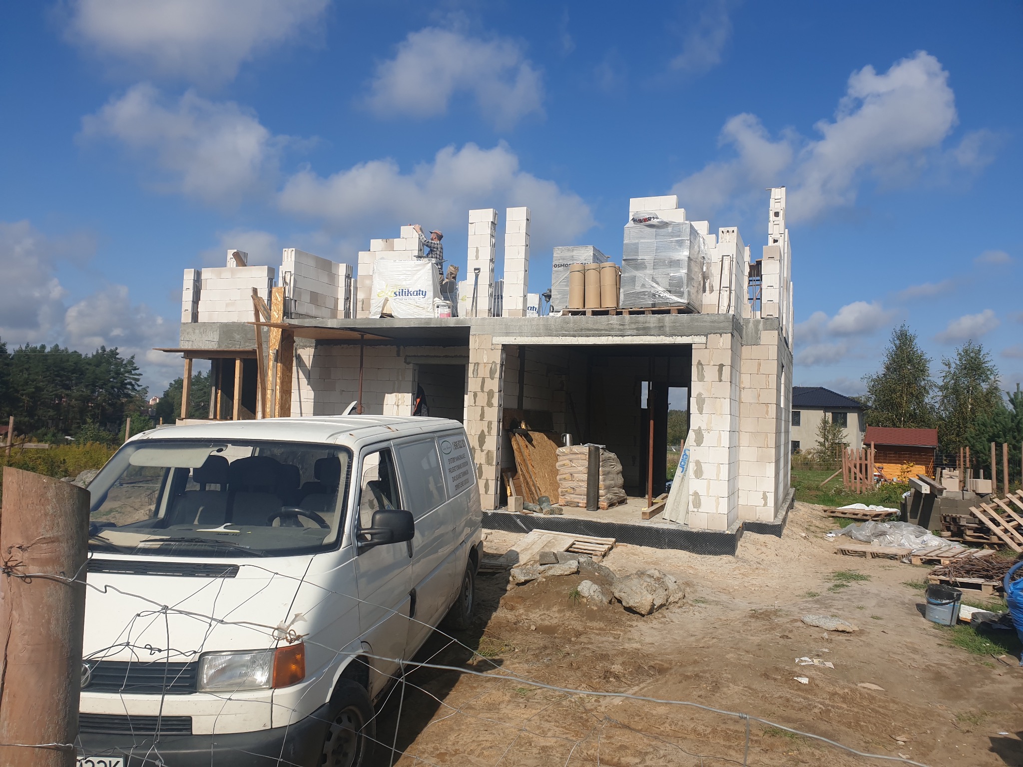 Wznoszenie ścian domu z białych bloczków silikatowych, widoczny częściowo załadowany materiałami budowlanymi biały samochód dostawczy na tle placu budowy i błękitnego nieba.