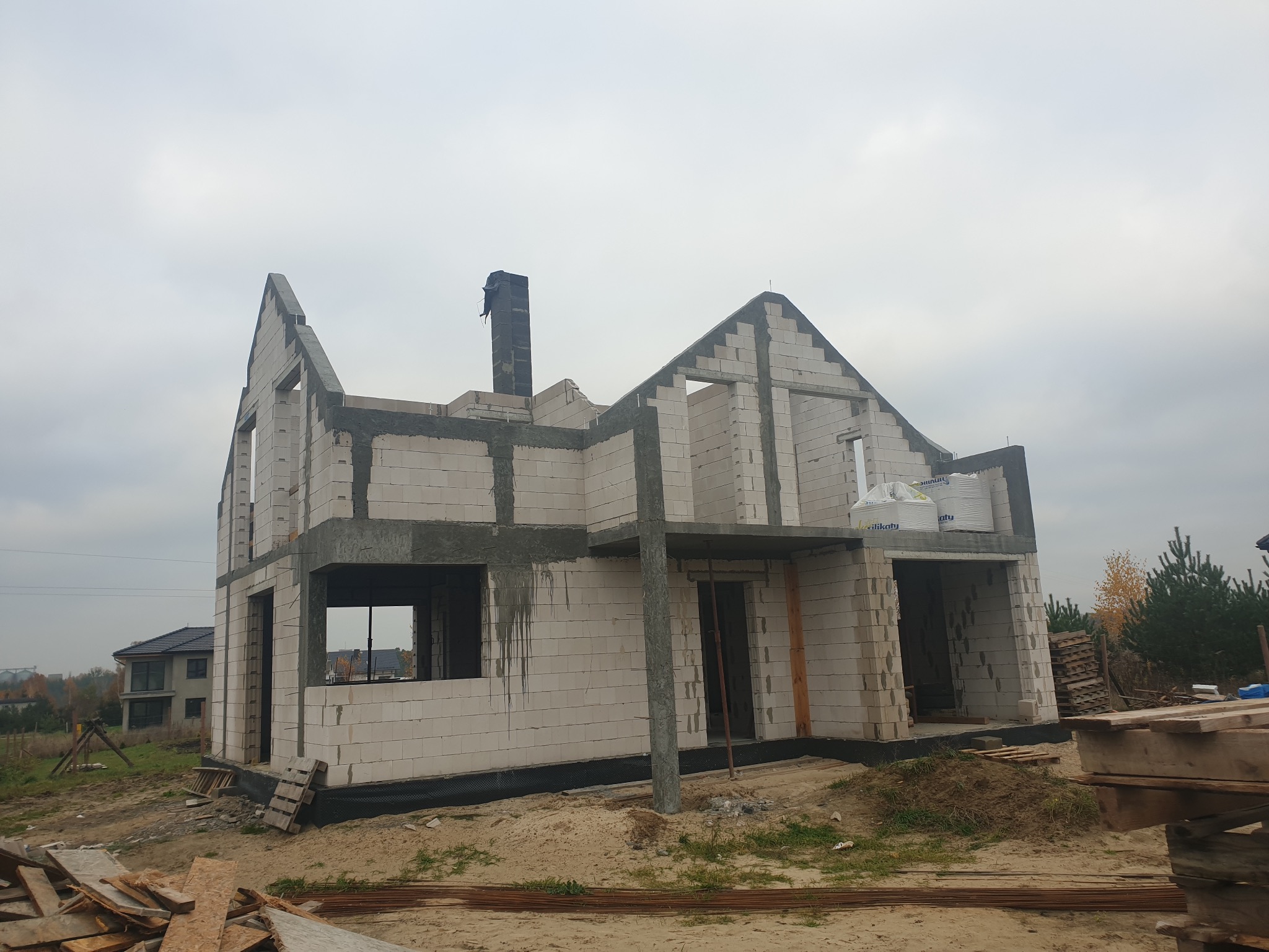 Surowy stan dwukondygnacyjnego budynku z białych bloczków, z widocznym kominem i otworami okiennymi, na tle pochmurnego nieba i drugiego domu w oddali.
