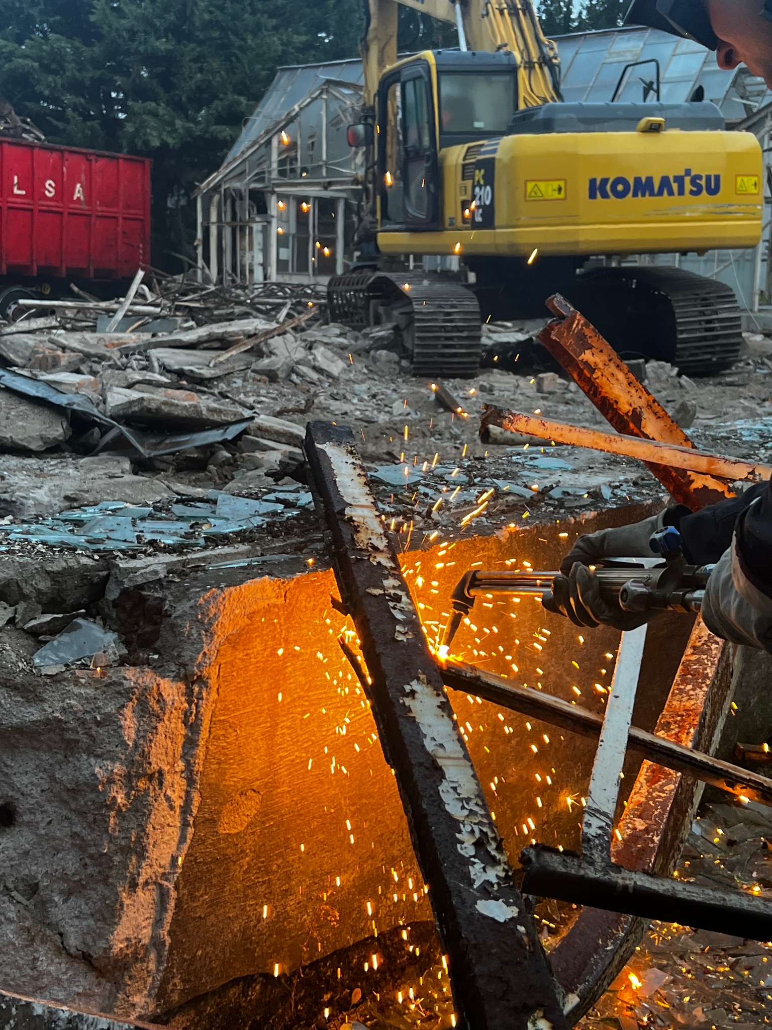 Demontaż konstrukcji stalowej palnikiem acetylenowo-tlenowym na tle zrujnowanego budynku i koparki Komatsu PC210, z widocznym kontenerem LSA w tle.