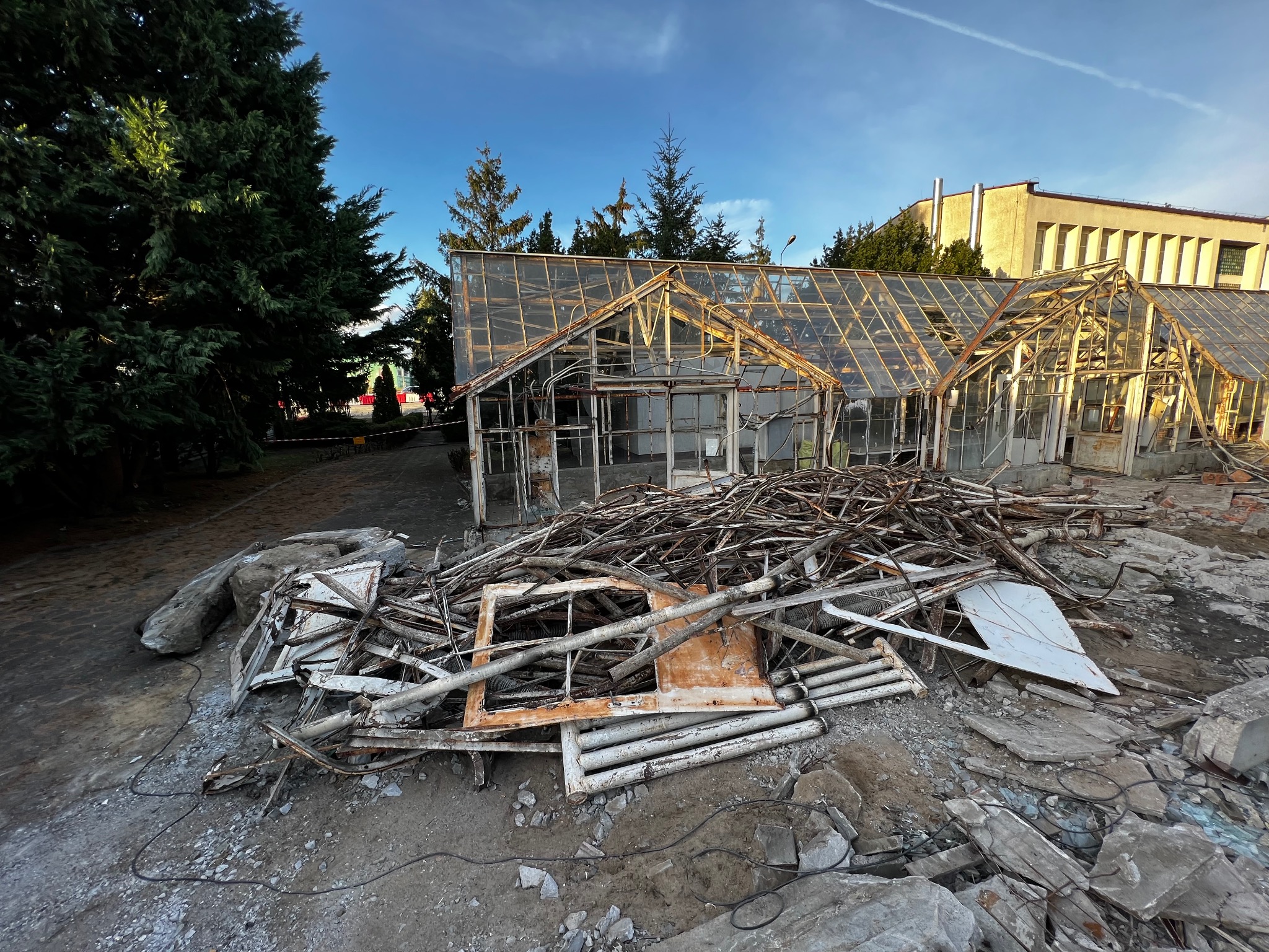 Zdemontowana, zniszczona konstrukcja szklarni z widocznymi elementami metalowymi i szklanymi, leżąca na ziemi w pobliżu budynku i drzew.