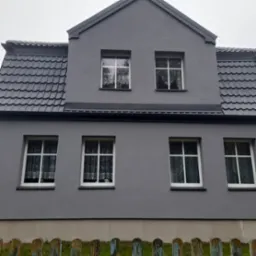 Szary dom dwupiętrowy z białymi oknami i ciemnoszarym dachem z dachówki, widziany zza starego, drewnianego płotu.