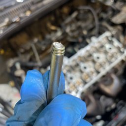 MB Rescue&Care - Zużyty zawór samochodowy w dłoni w niebieskiej rękawicy, z widocznymi nagarami i zabrudzeniami, na tle silnika samochodowego.