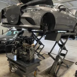 Szary samochód marki Mercedes na podnośniku warsztatowym, z zdjętym kołem i osłoną na masce, obok silnik na wózku w warsztacie samochodowym.